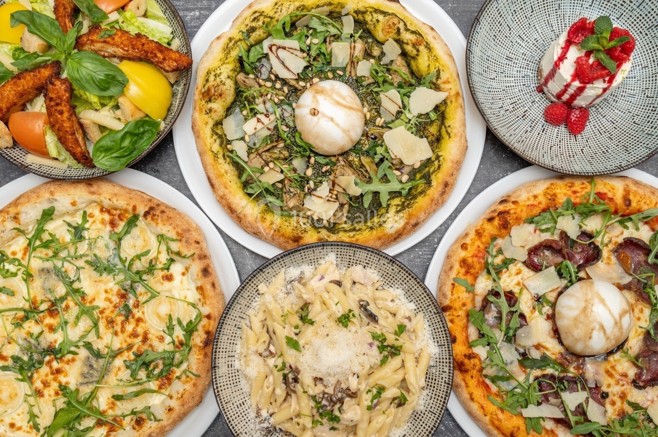 Assortiment de pizzas, pâtes et desserts présentés sur une table, garnis de légumes, fromages et herbes fraîches.