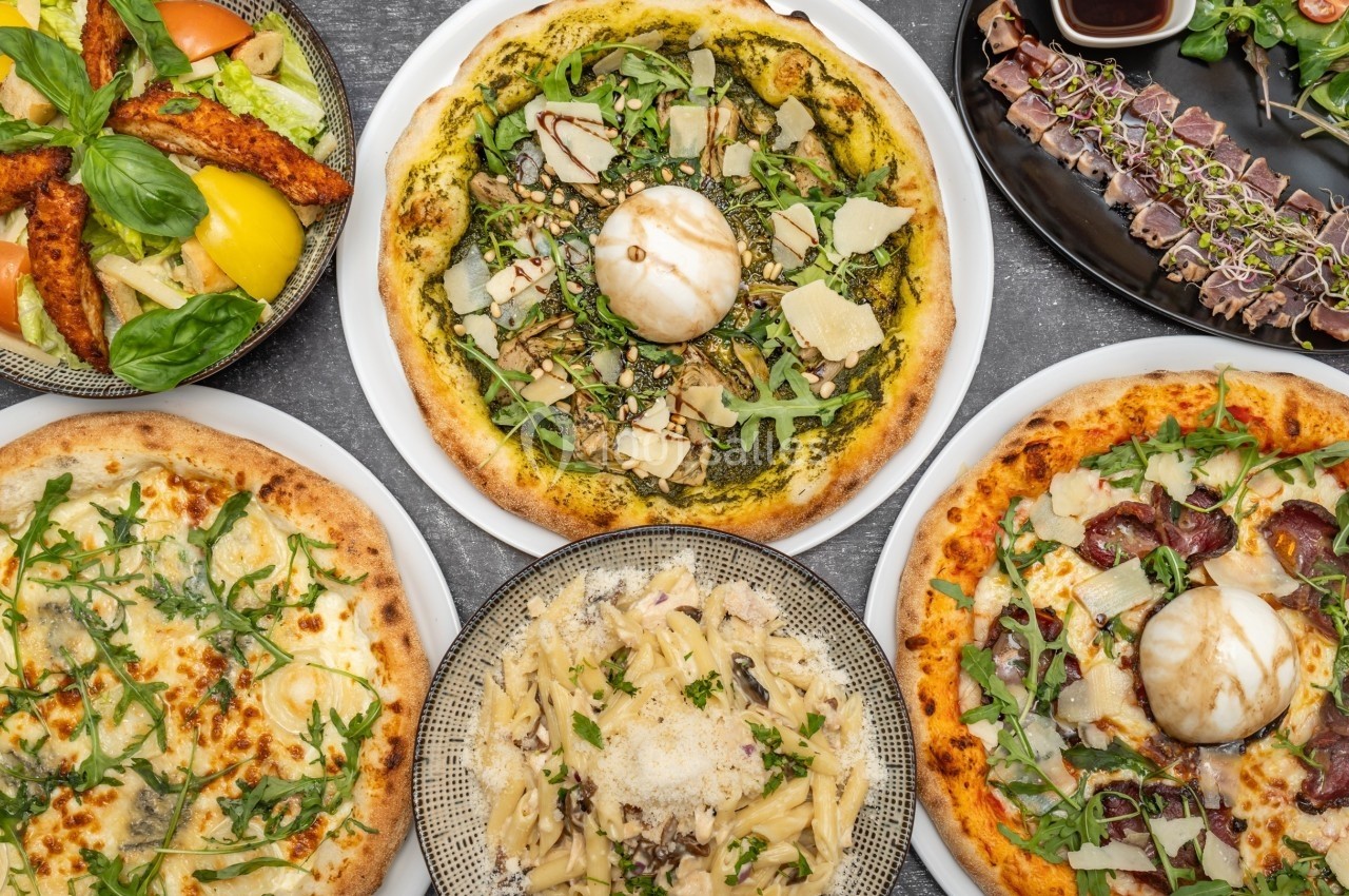 Assortiment de plats italiens comprenant pizzas, pâtes, salade et carpaccio, présenté sur une table grise.