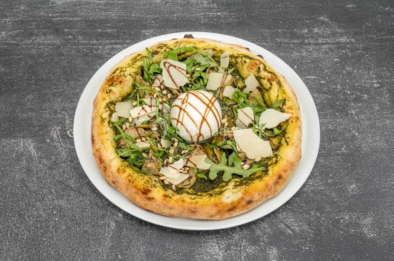 Pizza garnie de roquette, copeaux de parmesan, pesto et œuf poché, servie sur une assiette blanche.