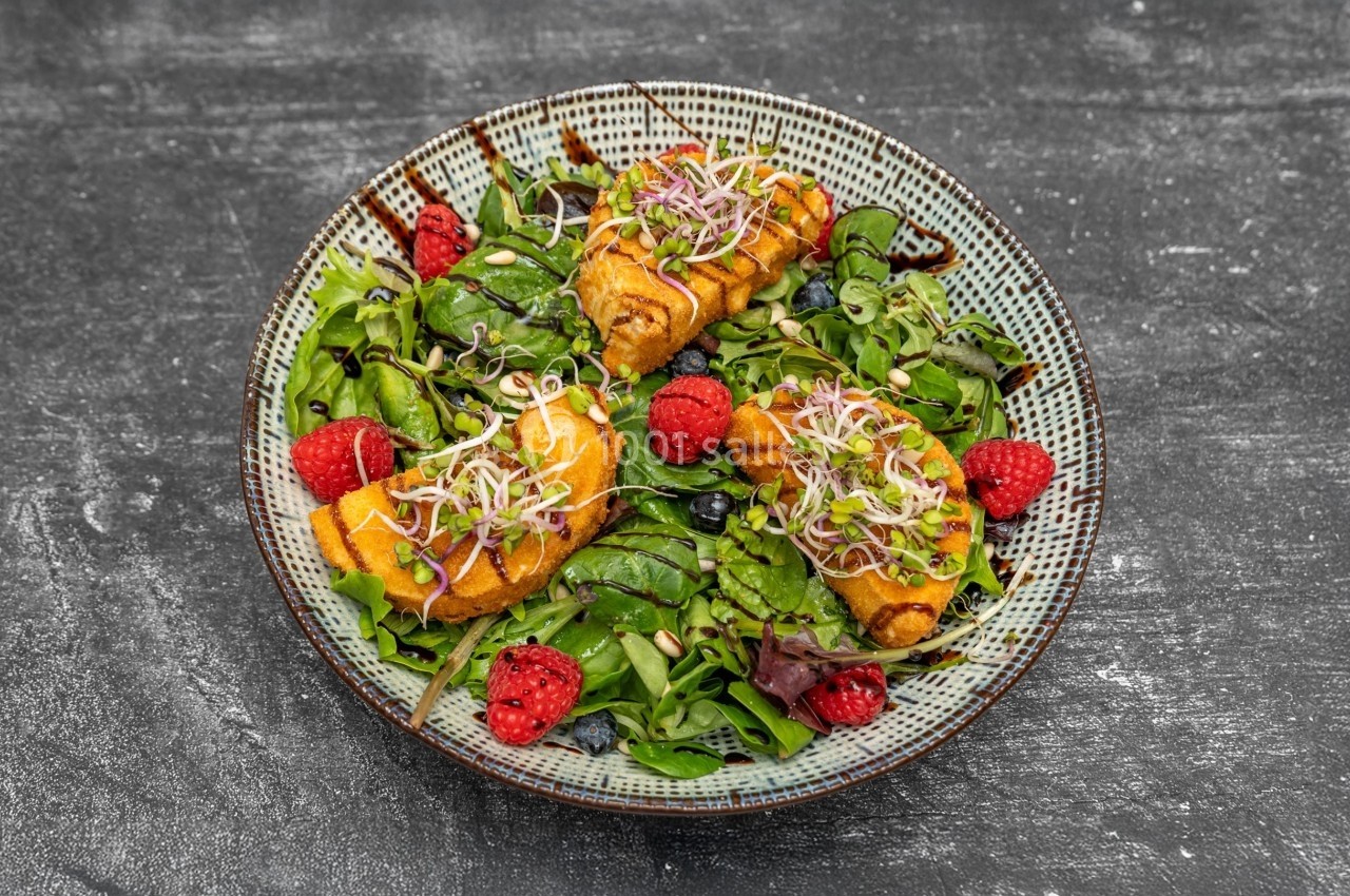 Assiette de salade verte garnie de framboises, pousses germées et rouleaux frits, sur une table sombre.