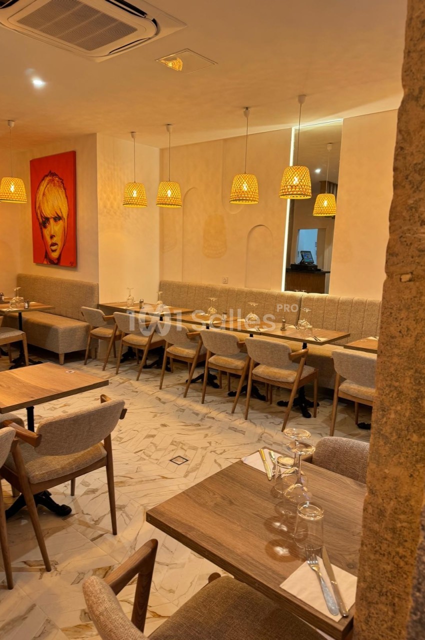 Salle de restaurant chaleureuse avec tables en bois, chaises rembourrées, luminaires suspendus et tableau mural coloré.