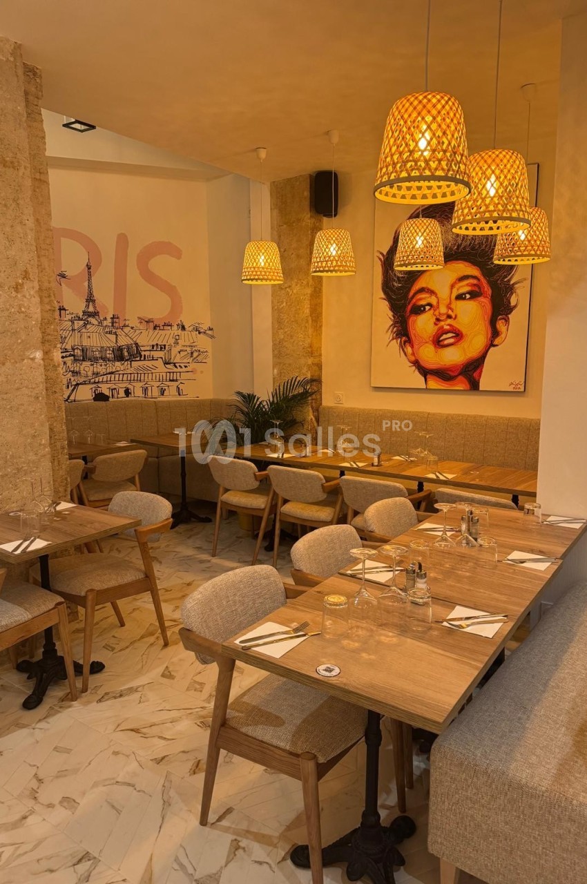 Salle de restaurant chaleureuse avec tables en bois, chaises rembourrées, luminaires suspendus et tableau coloré au mur.