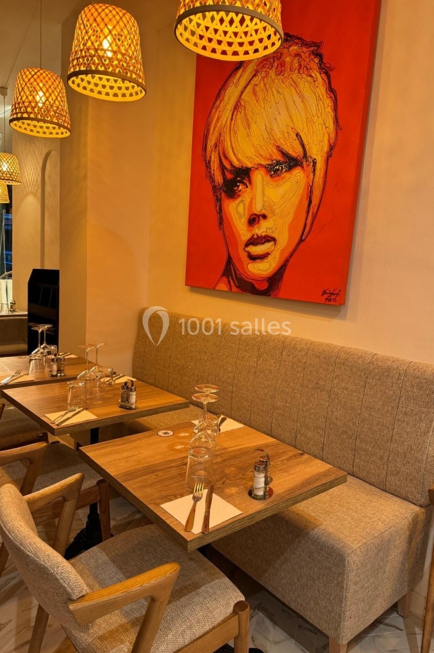 Salle de restaurant avec tables en bois, chaises rembourrées, luminaires suspendus et un tableau coloré au mur.
