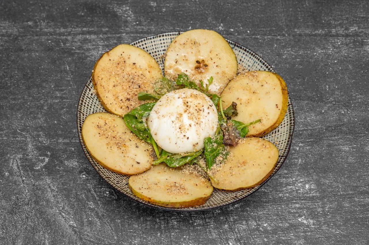 Assiette garnie de tranches de pommes de terre, burrata, roquette et assaisonnement sur une table sombre.