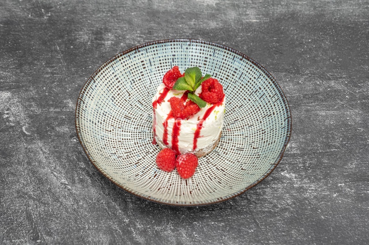 Dessert à base de mousse blanche, décoré de framboises, coulis rouge et une feuille de menthe, présenté dans une assiette.