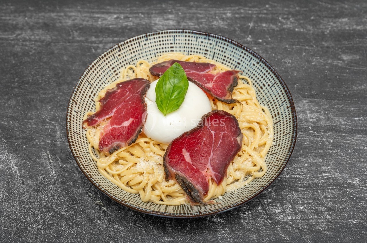 Assiette de pâtes garnie de tranches de viande séchée, burrata et feuille de basilic, sur fond noir.