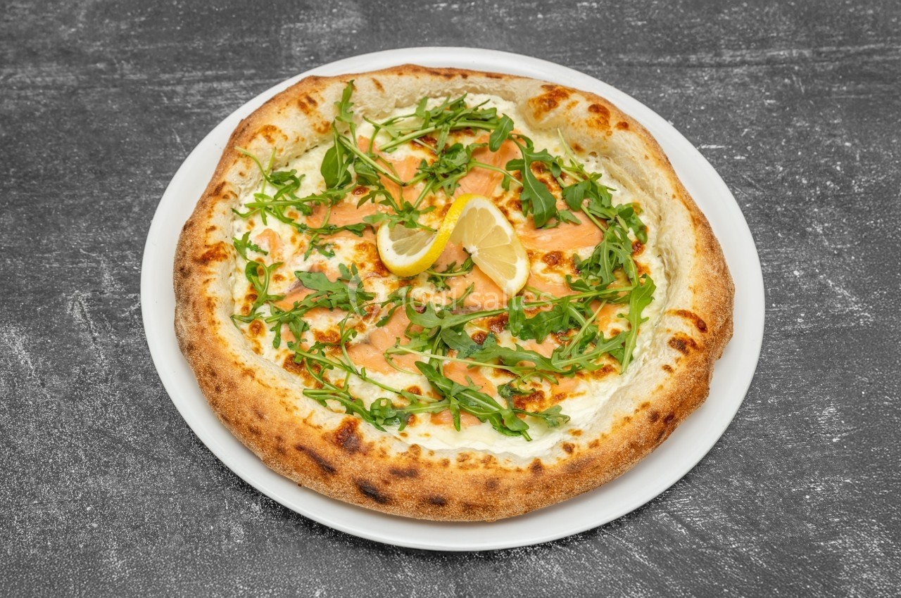 Pizza garnie de saumon fumé, roquette, citron et fromage, servie sur une assiette blanche sur fond gris.