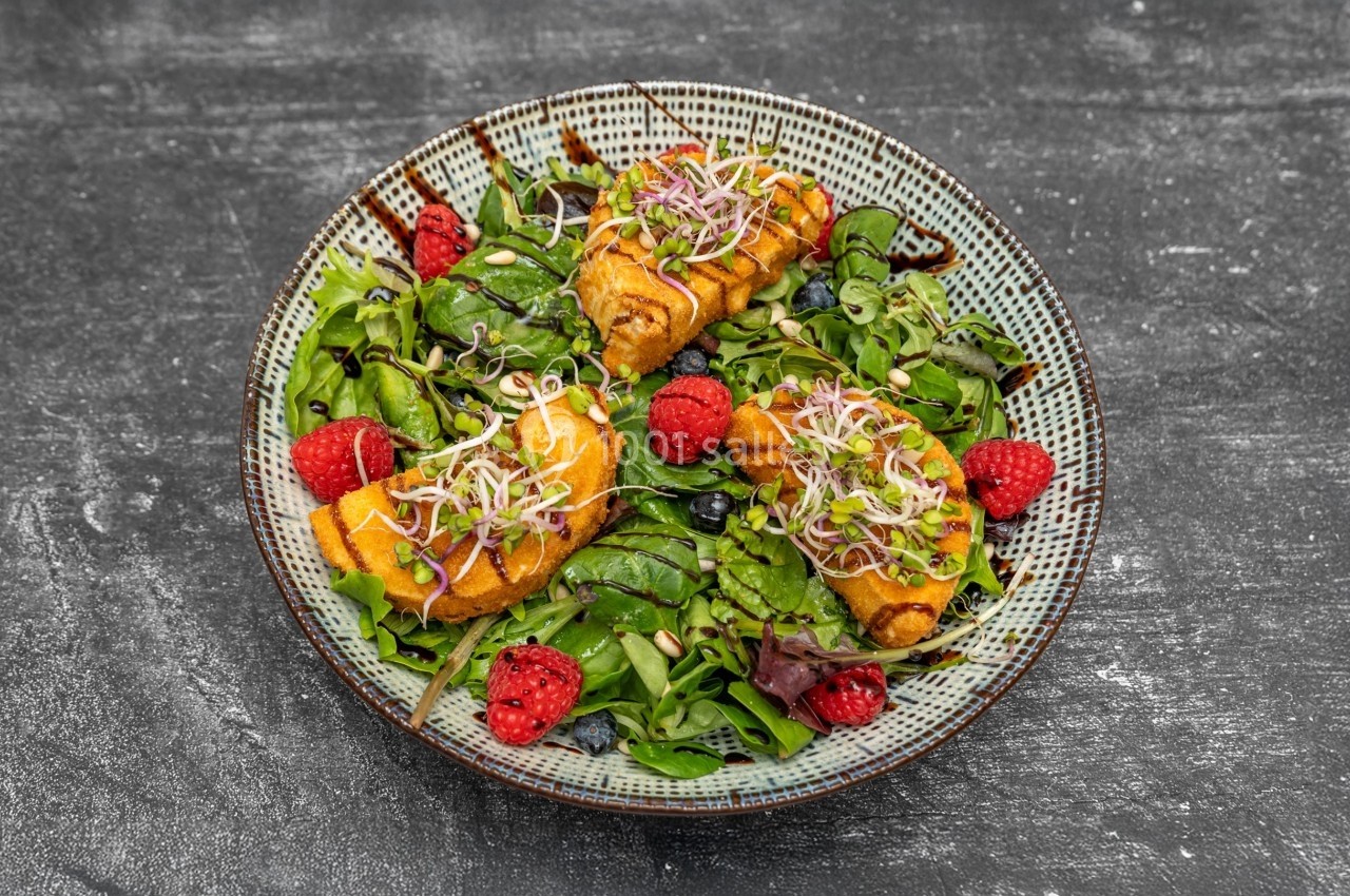Assiette de salade verte garnie de framboises, pousses germées et rouleaux croustillants sur une table sombre.