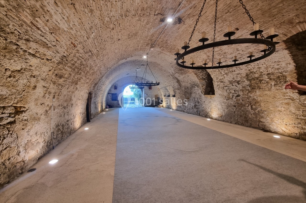 Voûte en pierre éclairée avec des lustres en fer forgé, menant à une ouverture lumineuse au fond.