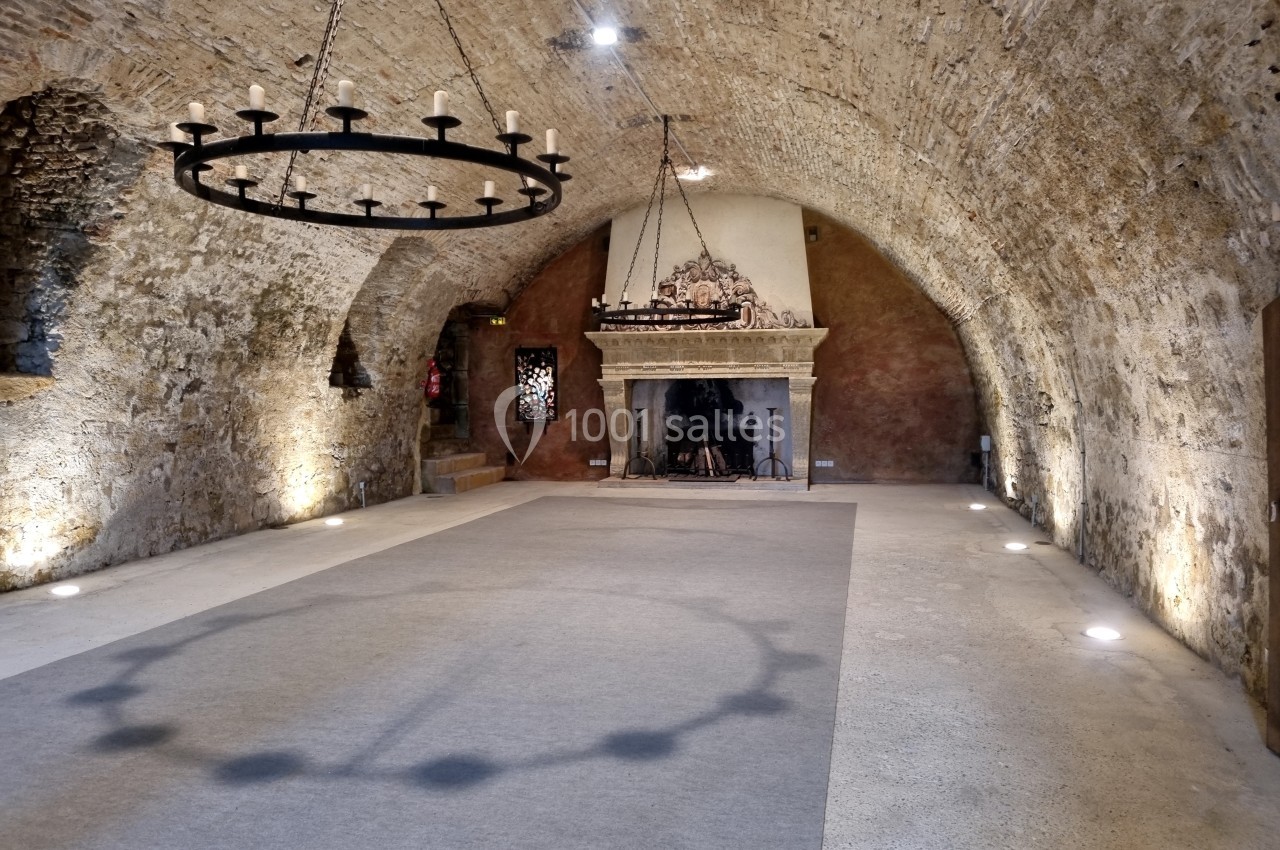 Salle voûtée en pierre avec un lustre en fer forgé, une cheminée ancienne et un éclairage au sol.