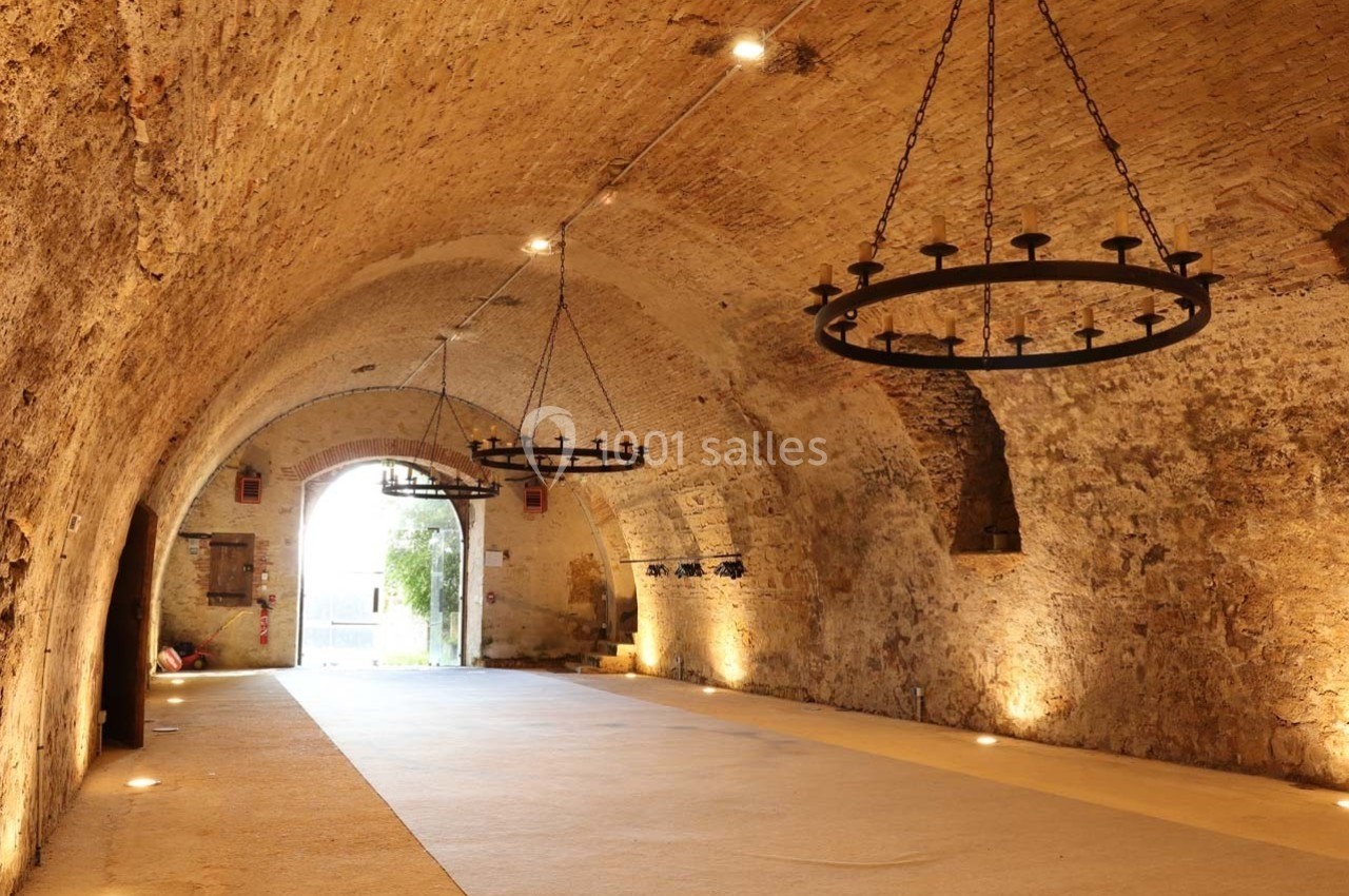 Salle voûtée en pierre avec chandeliers suspendus, éclairage chaleureux et ouverture donnant sur l'extérieur.