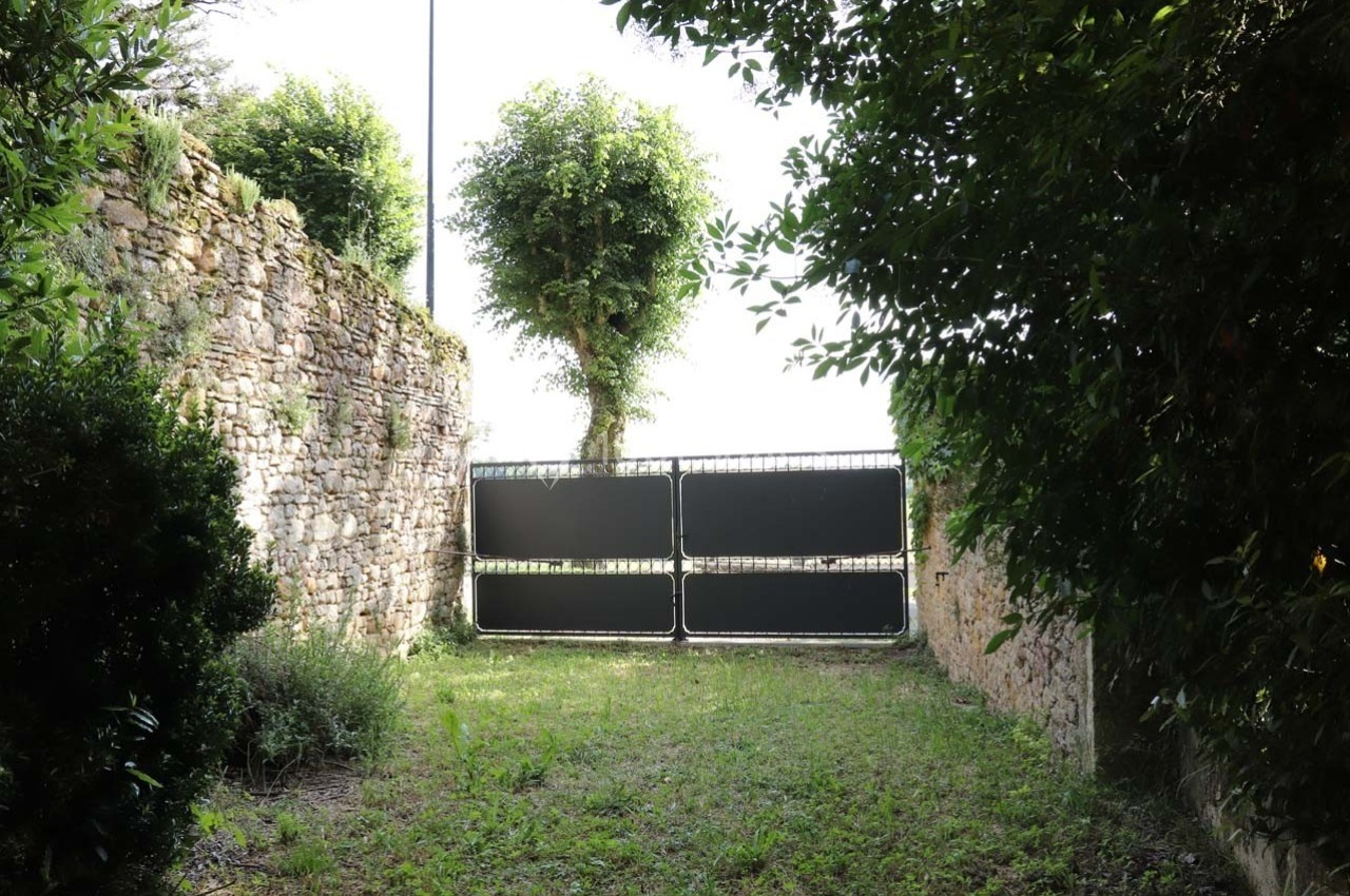 Portail métallique noir fermé entre deux murs en pierre, entouré de végétation et d'arbustes.