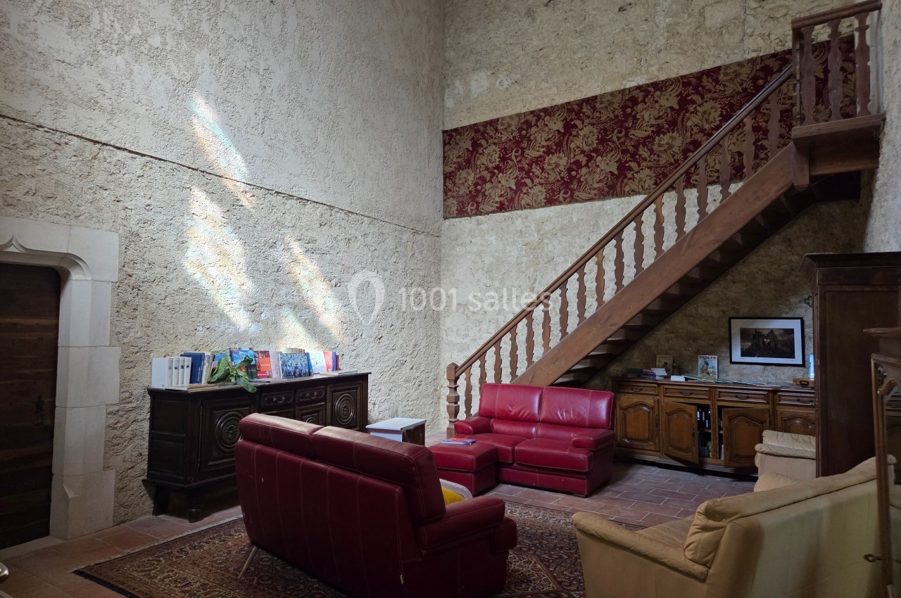 Salon avec murs en pierre, canapé rouge, escalier en bois, tapis au sol et meubles anciens en bois.