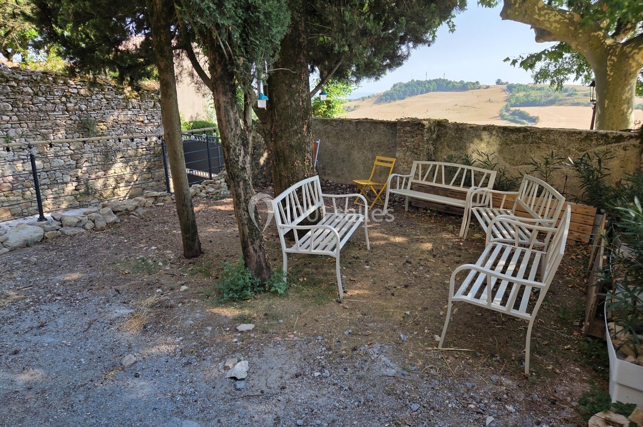 Bancs en métal blanc disposés sous des arbres dans une cour ensoleillée avec vue sur un paysage rural.
