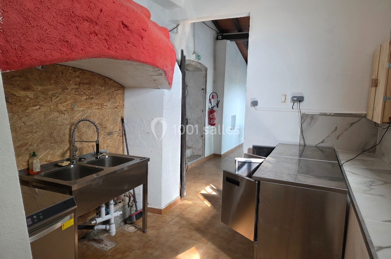 Cuisine équipée avec évier double, plans de travail en inox, lave-vaisselle et murs blancs dans un espace lumineux.