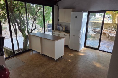 Cuisine lumineuse avec îlot central, réfrigérateur blanc et grandes baies vitrées donnant sur un espace extérieur arboré.