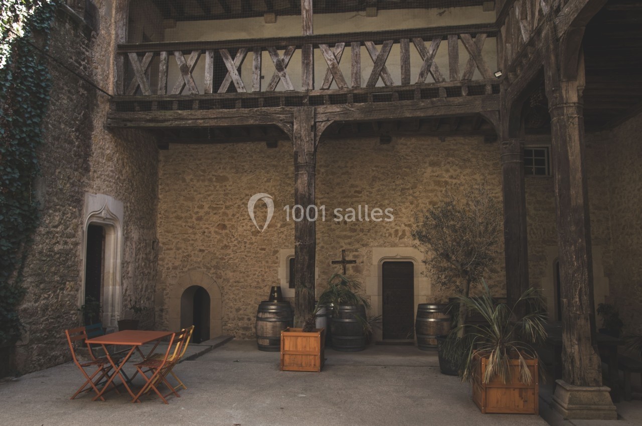 Cour intérieure en pierre avec des poutres en bois, des tonneaux, des plantes et une table avec des chaises orange.
