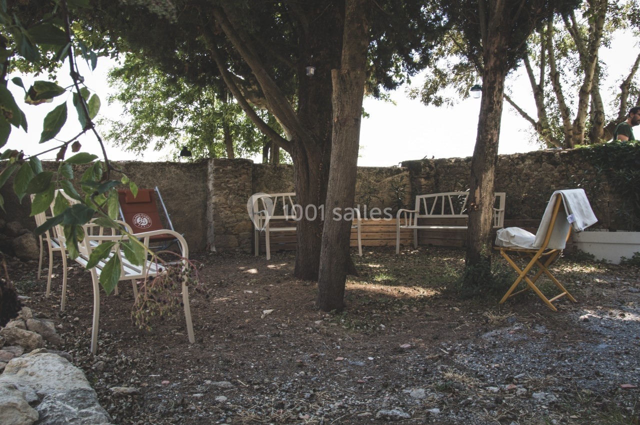 Chaises et bancs disposés sous des arbres dans un jardin ombragé entouré de murs en pierre.