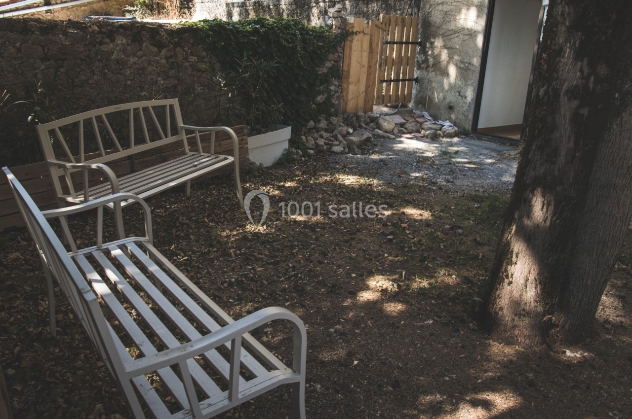 Deux bancs blancs en métal placés dans un jardin ombragé avec un arbre, un mur en pierre et une clôture en bois.