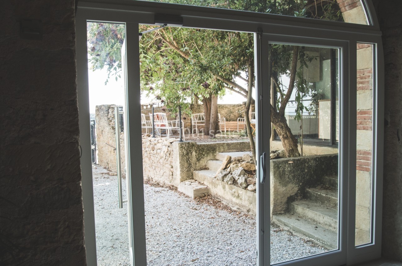 Vue depuis une porte vitrée donnant sur une cour extérieure avec arbres, murs en pierre et mobilier de jardin.