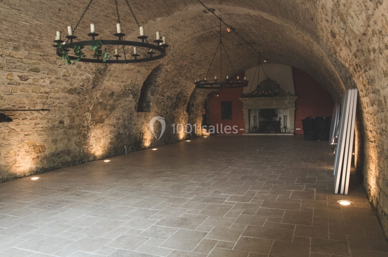 Salle voûtée en pierre avec un sol carrelé, éclairage tamisé et cheminée au fond.