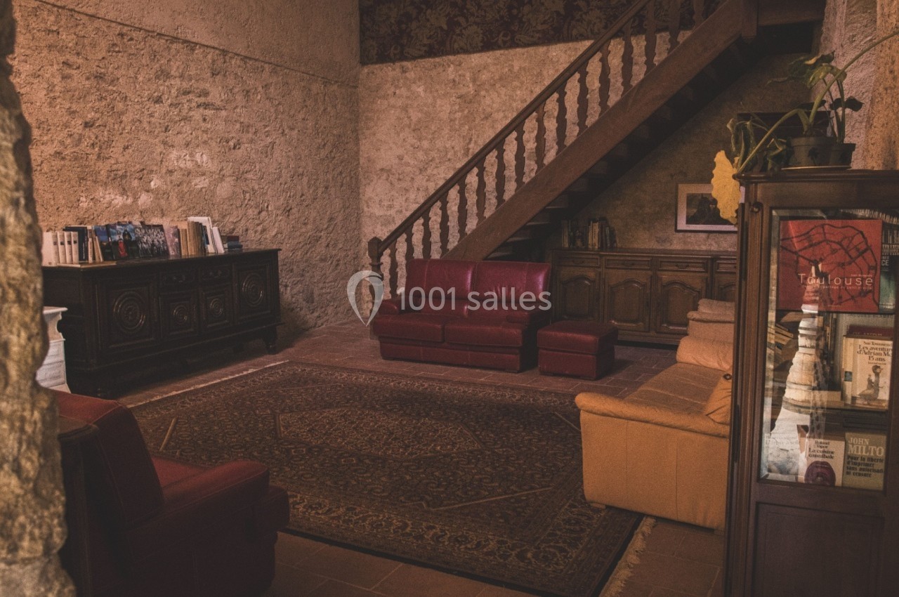 Salon rustique avec murs en pierre, canapé rouge et beige, tapis oriental, escalier en bois et meubles anciens.