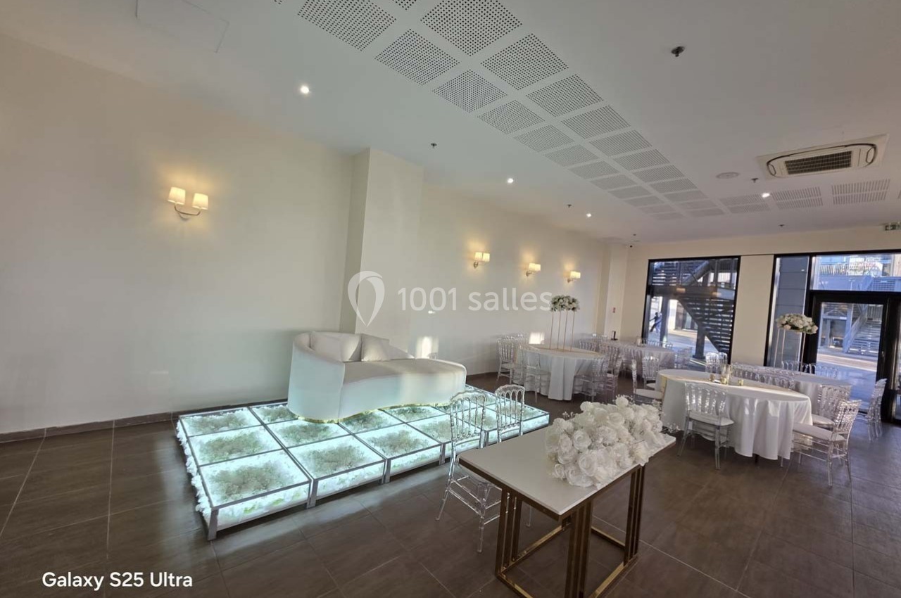 Salle de réception moderne avec un canapé blanc sur une plateforme lumineuse, tables décorées et grandes fenêtres.