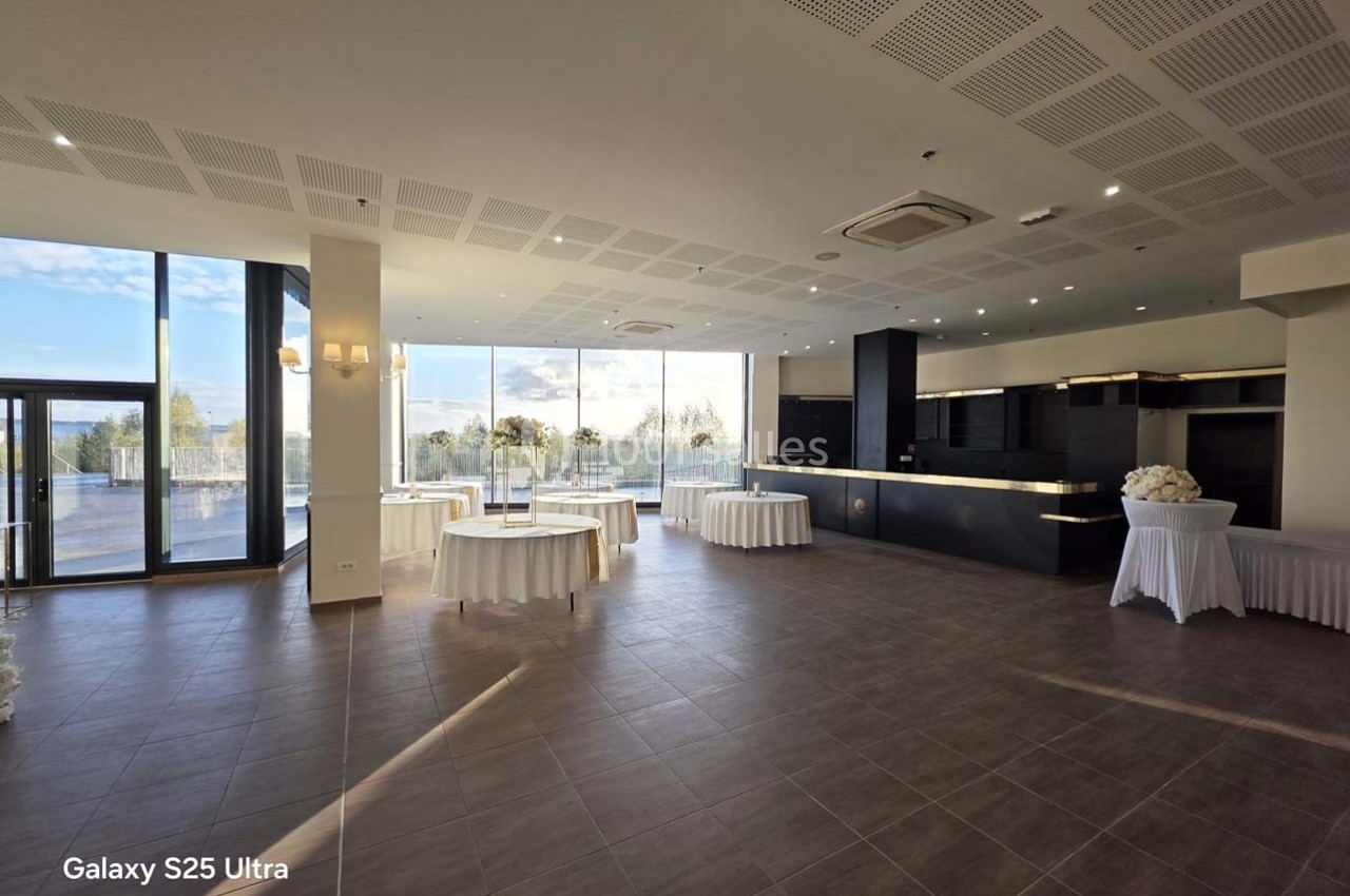 Salle lumineuse avec grandes baies vitrées, tables rondes drapées de blanc et comptoir moderne en arrière-plan.