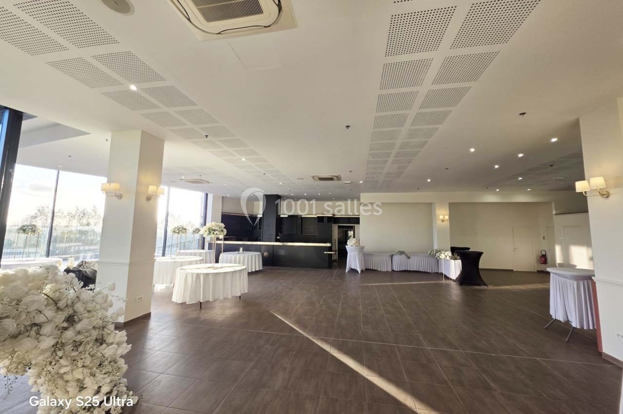 Salle de réception lumineuse avec grandes baies vitrées, tables décorées de fleurs blanches et sol en carrelage foncé.