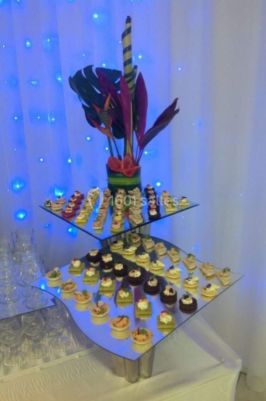 Plateau de canapés variés disposés sur une structure à trois niveaux, décorée de feuilles et fleurs tropicales.