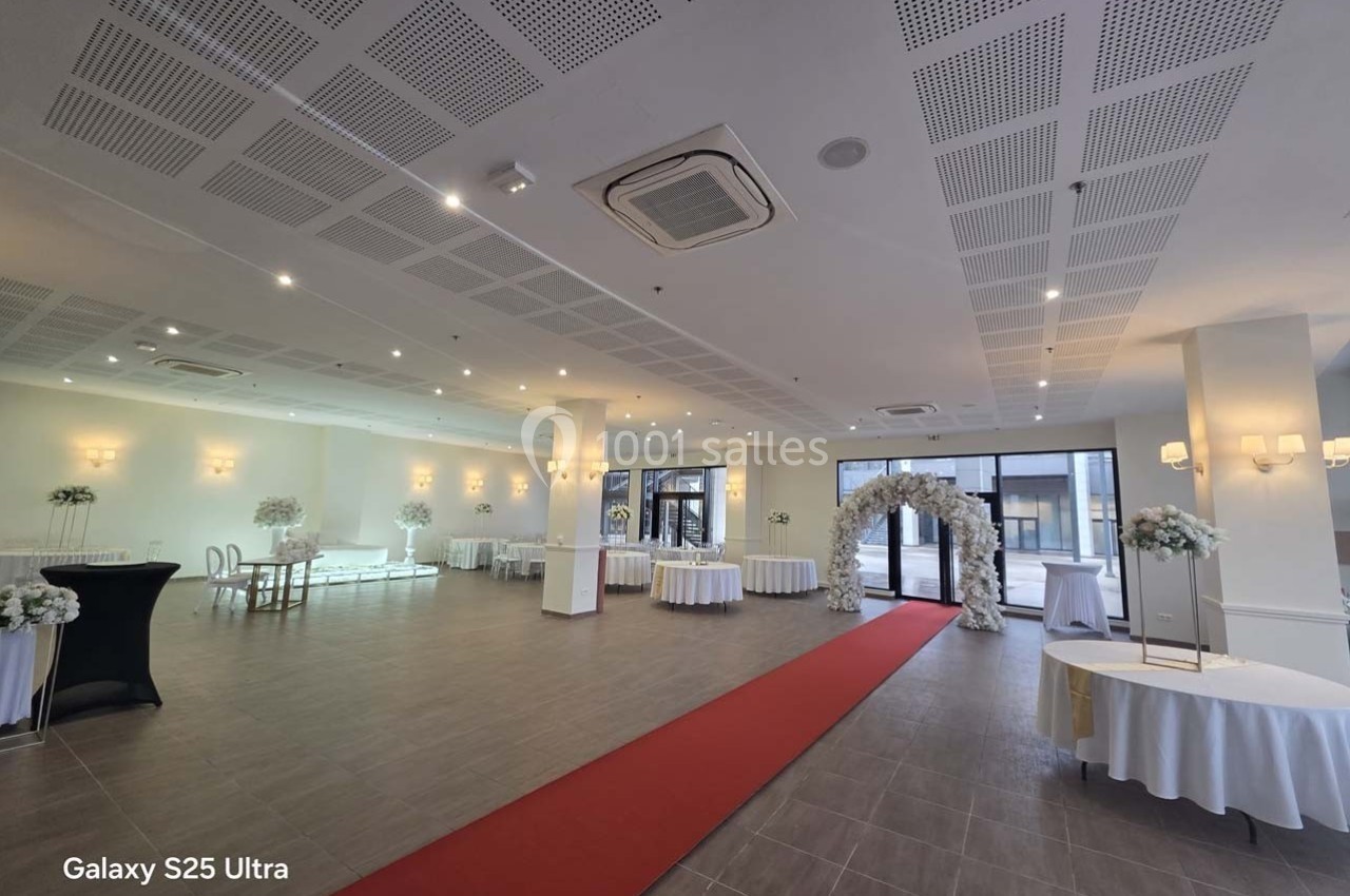 Salle de réception lumineuse avec tables décorées, arche florale et tapis rouge menant à l'entrée.