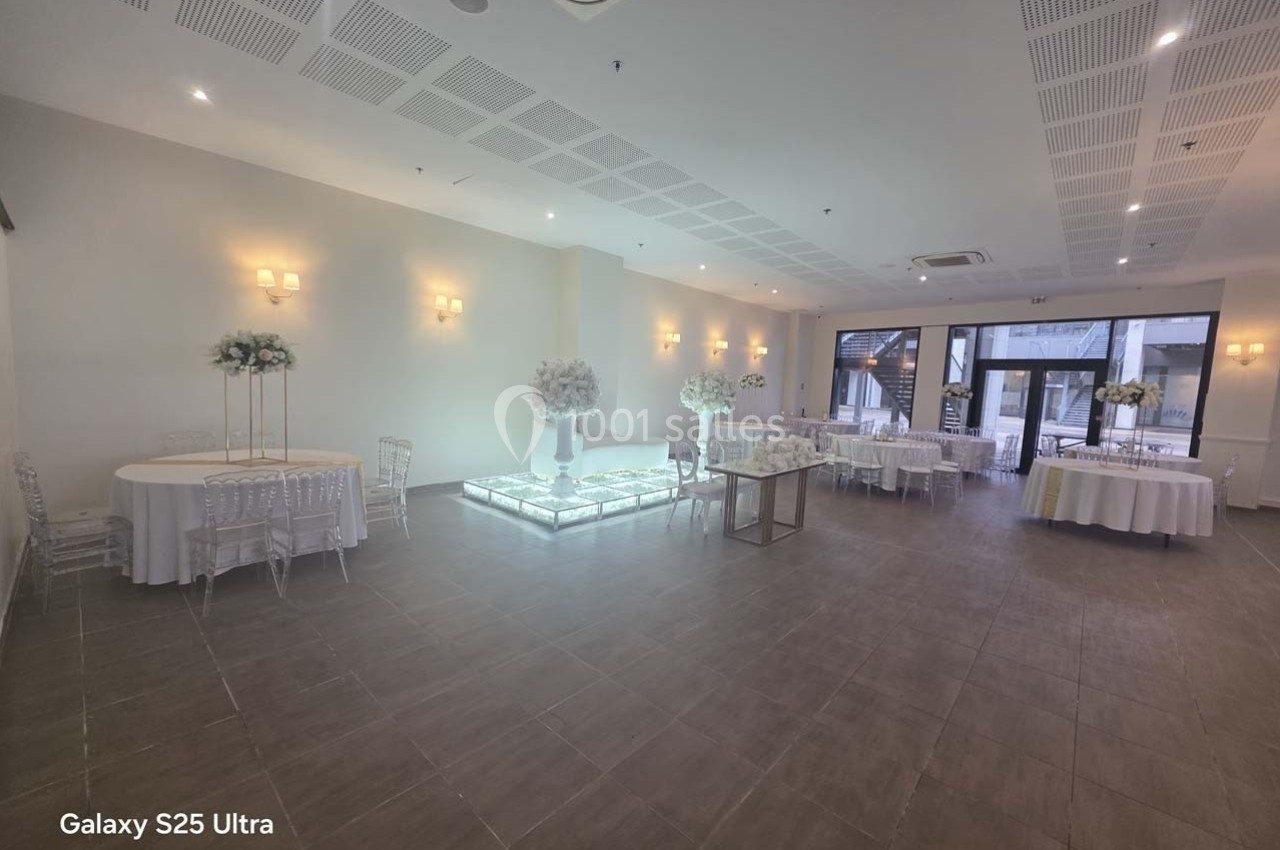 Salle de réception lumineuse avec tables décorées de fleurs blanches, sol carrelé et grandes baies vitrées.