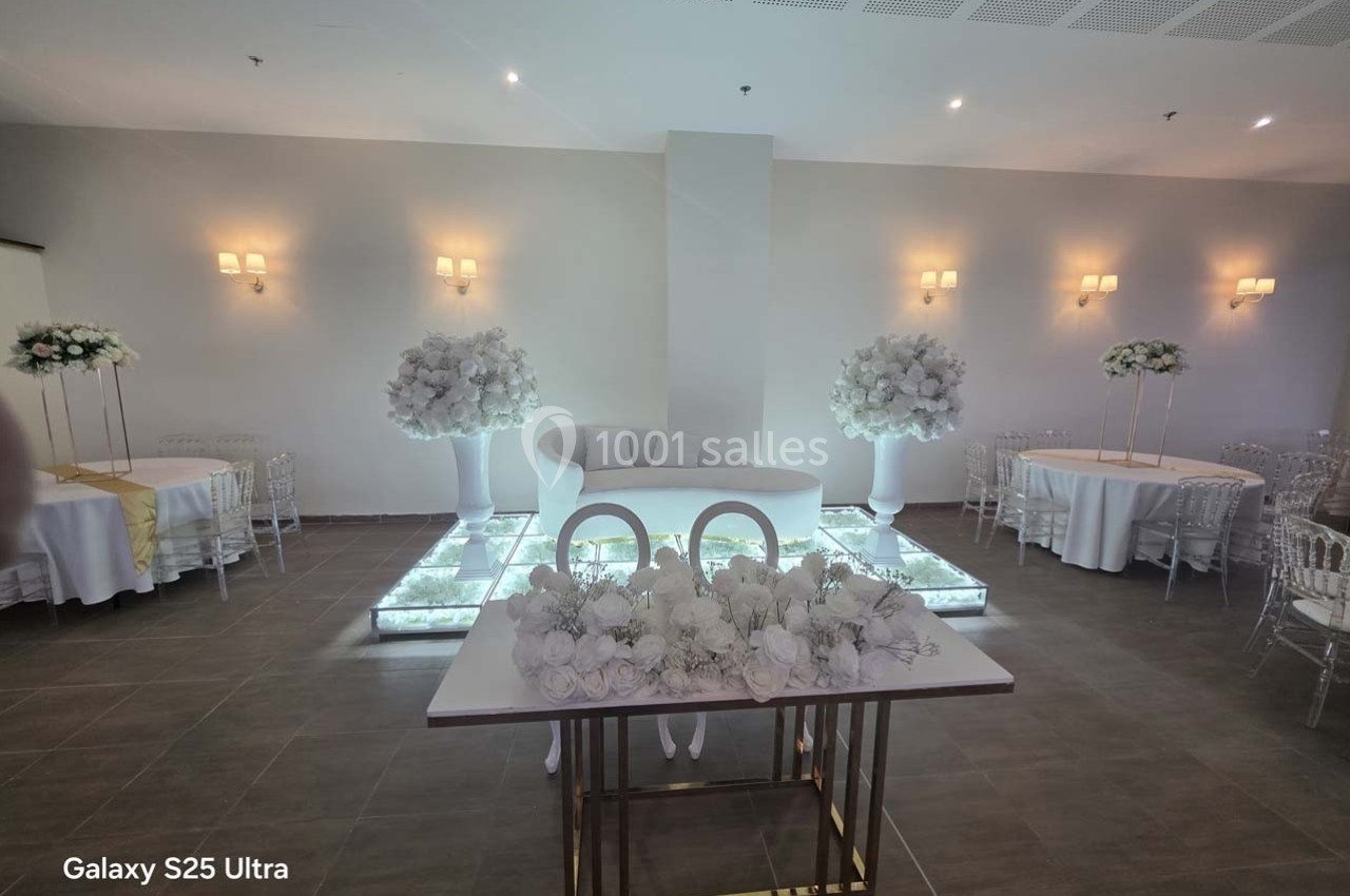 Salle de réception décorée avec des fleurs blanches, des tables rondes et un canapé sur une estrade lumineuse.