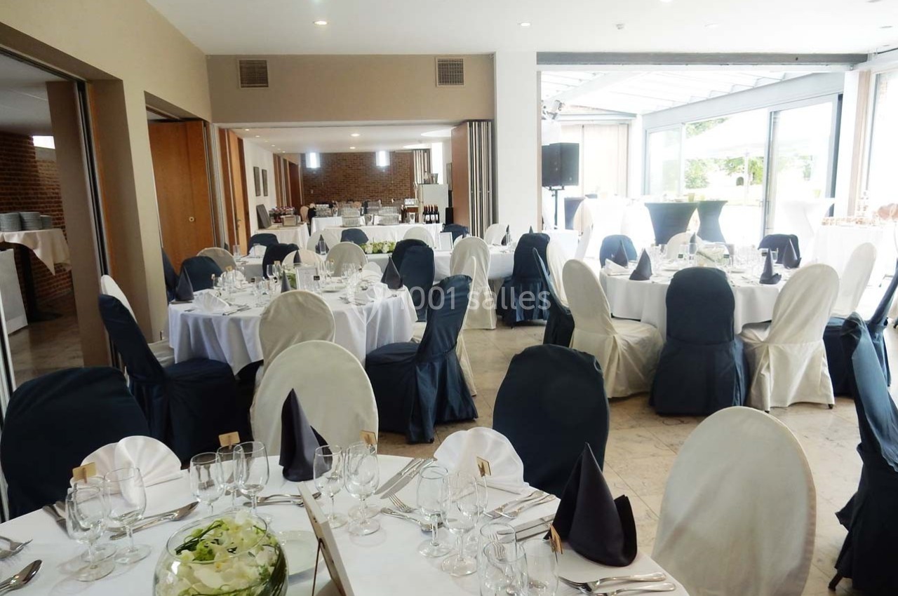 Salle de réception lumineuse avec des tables rondes dressées, nappes blanches et chaises couvertes.