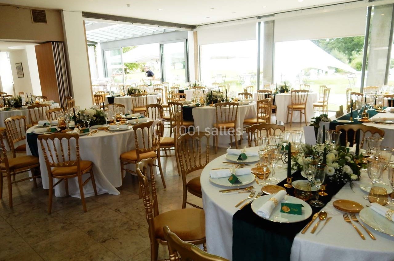 Salle de réception décorée avec des tables rondes dressées, nappes blanches, chaises dorées et arrangements floraux.