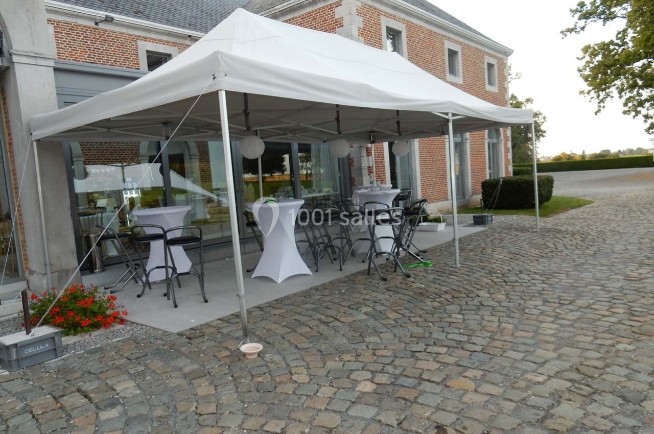 Tente blanche installée sur une terrasse pavée avec tables hautes, chaises et décorations pour un événement extérieur.