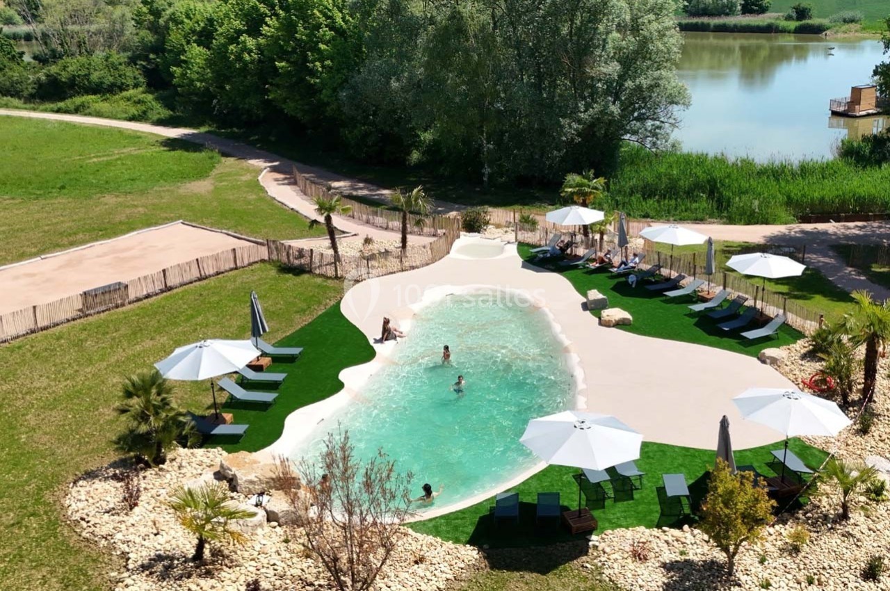 Vue aérienne d'une piscine naturelle entourée de transats, parasols, pelouse et végétation, près d'un lac.