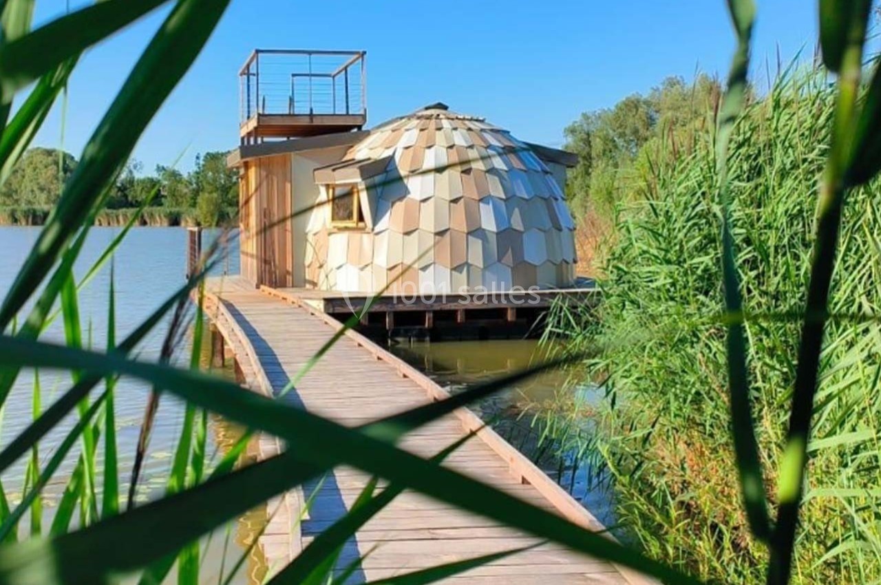 Cabane en bois au design géométrique sur pilotis, reliée à une passerelle au bord d’un lac entouré de roseaux.
