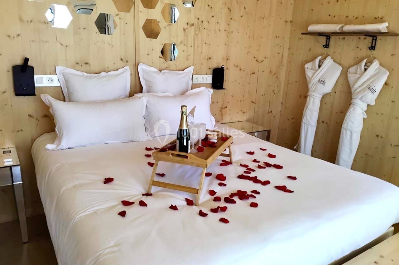 Lit double avec draps blancs, pétales de rose, plateau avec champagne et peignoirs accrochés sur un mur en bois.