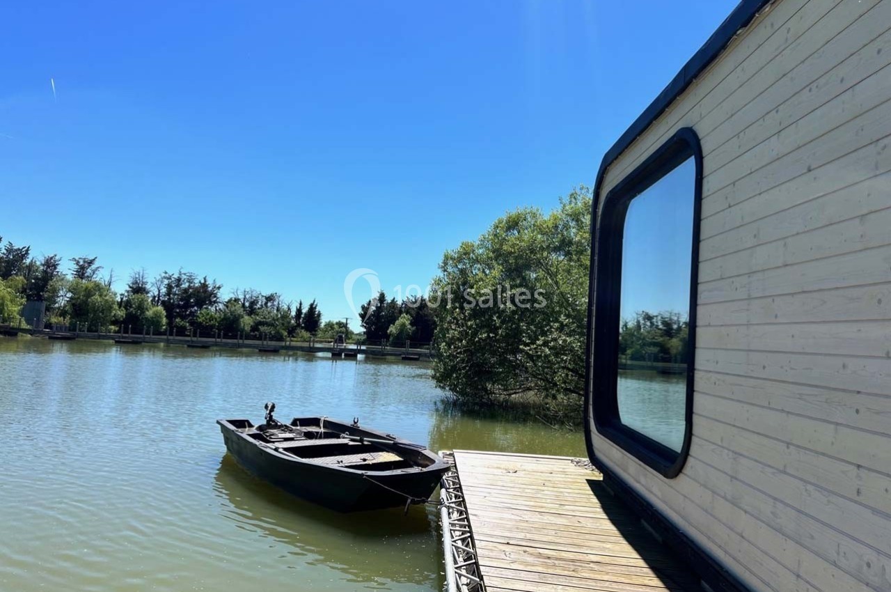 Une petite barque noire amarrée près d'une cabane en bois sur un lac entouré de végétation sous un ciel bleu.