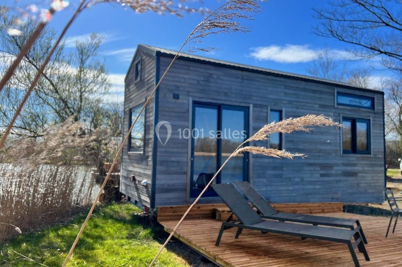 Petite maison en bois avec terrasse, entourée de végétation, près d'un plan d'eau sous un ciel bleu.