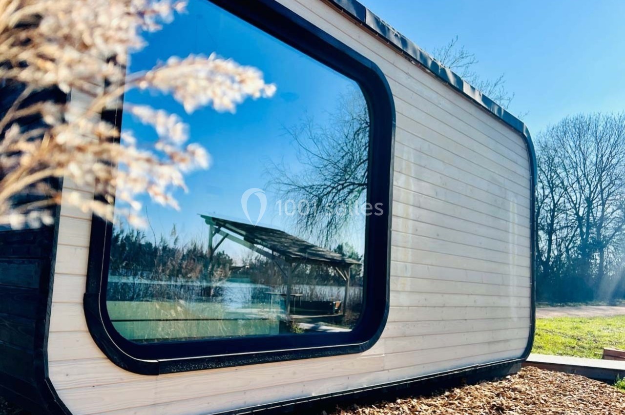 Cabane moderne en bois avec une grande baie vitrée, entourée de nature et éclairée par un ciel ensoleillé.