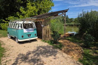 Espace extérieur avec fauteuils en bois, table ronde, et un van rétro bleu stationné en arrière-plan.