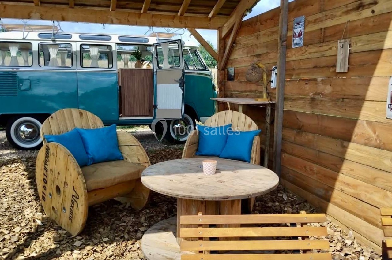 Espace extérieur avec fauteuils en bois, table ronde, et un van rétro bleu stationné en arrière-plan.