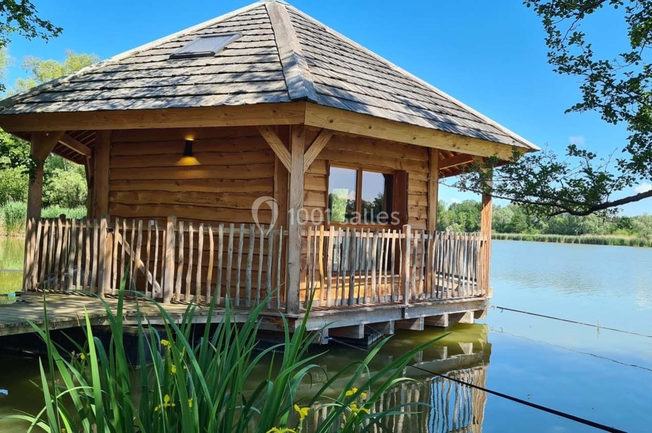 Cabane en bois sur pilotis au bord d'un étang, entourée de végétation et éclairée par un ciel bleu.
