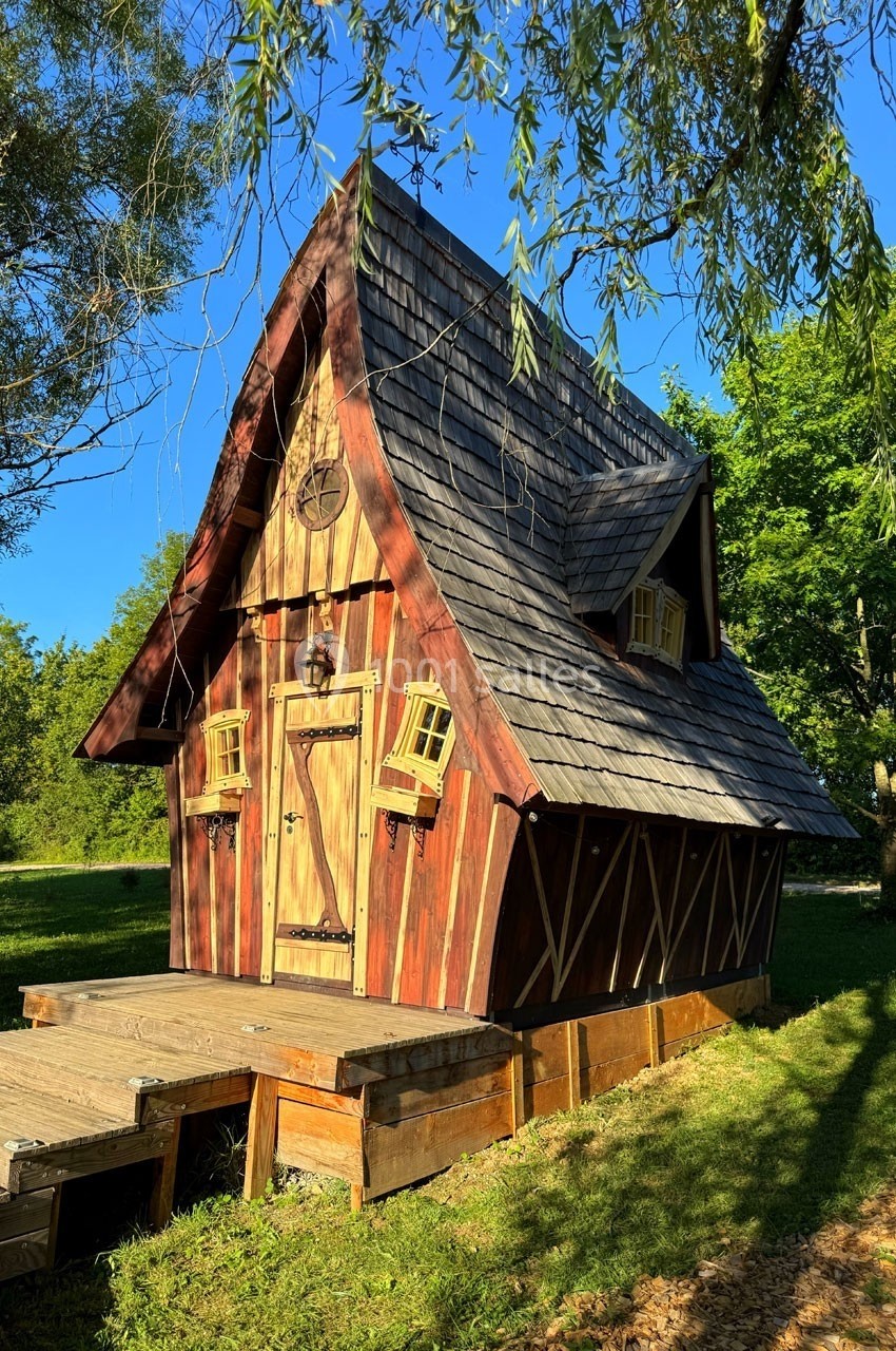 Cabane en bois au toit pentu, entourée d'arbres, éclairée par une lumière naturelle sous un ciel bleu.
