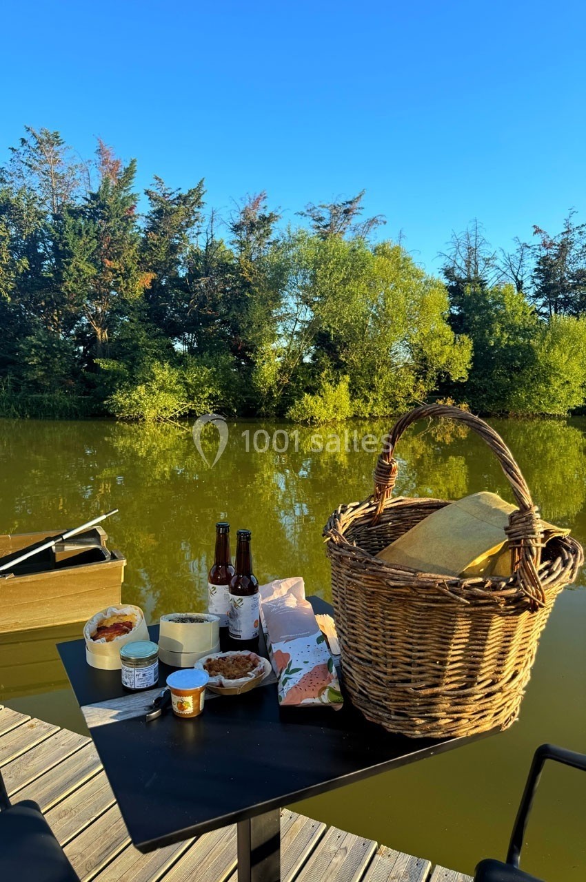 Un panier en osier et des produits alimentaires sont disposés sur une table en bois près d'un étang entouré de végétation.