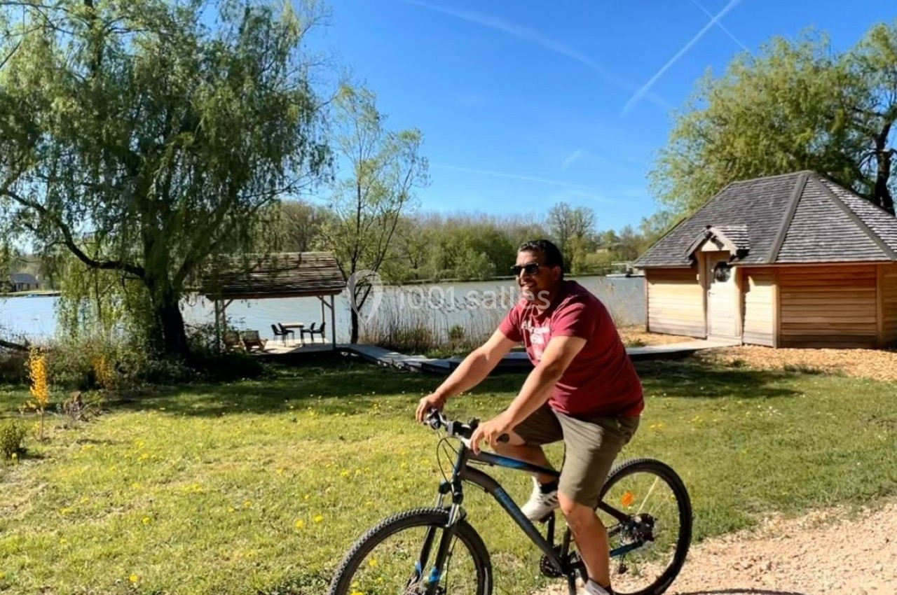 Un homme fait du vélo sur un chemin près d'un lac, entouré d'arbres et de petites constructions en bois.