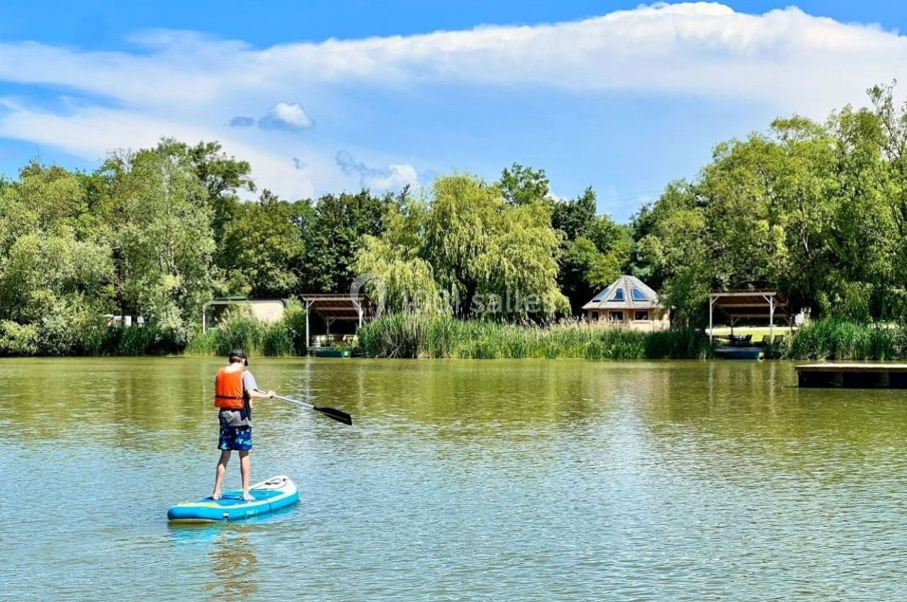 Un homme en gilet de sauvetage fait du paddle sur un lac entouré de végétation et de petites cabanes.