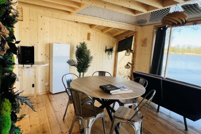 Intérieur d'une cabane en bois avec table ronde, chaises métalliques, canapé et terrasse extérieure visible.